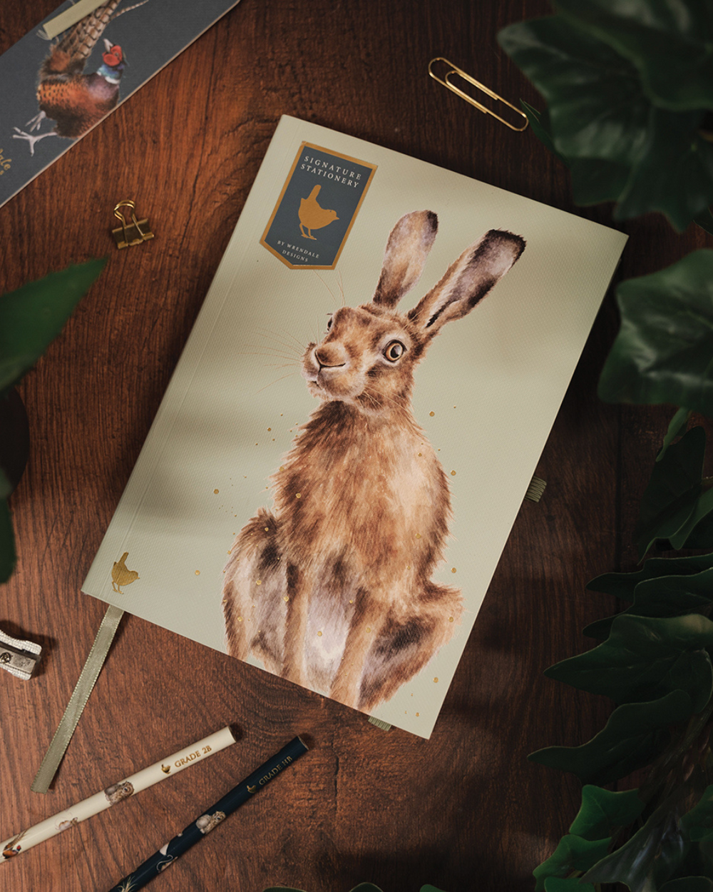 A5 Notizbuch „Hase“ aus der Signature Stationery Kollektion von Wrendale Designs, geschlossen auf Holztisch mit Pflanzen und passenden Bleistiften arrangiert.