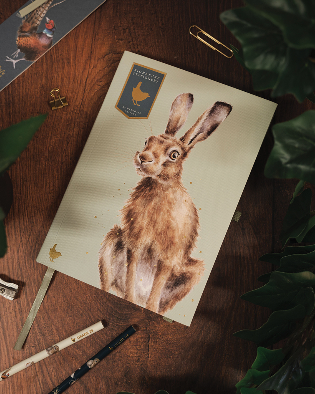 A5 Notizbuch „Hase“ aus der Signature Stationery Kollektion von Wrendale Designs, geschlossen auf Holztisch mit Pflanzen und passenden Bleistiften arrangiert.