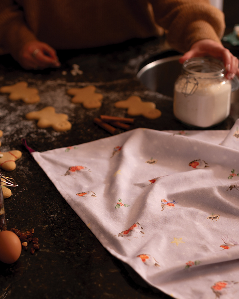 Ein weihnachtliches Geschirrtuch mit Rotkehlchen-Motiv liegt auf einer dunklen Küchenarbeitsfläche. Daneben stehen Zutaten wie Eier, Zimtstangen und ein Glas mit Mehl, im Hintergrund werden Plätzchen ausgestochen.