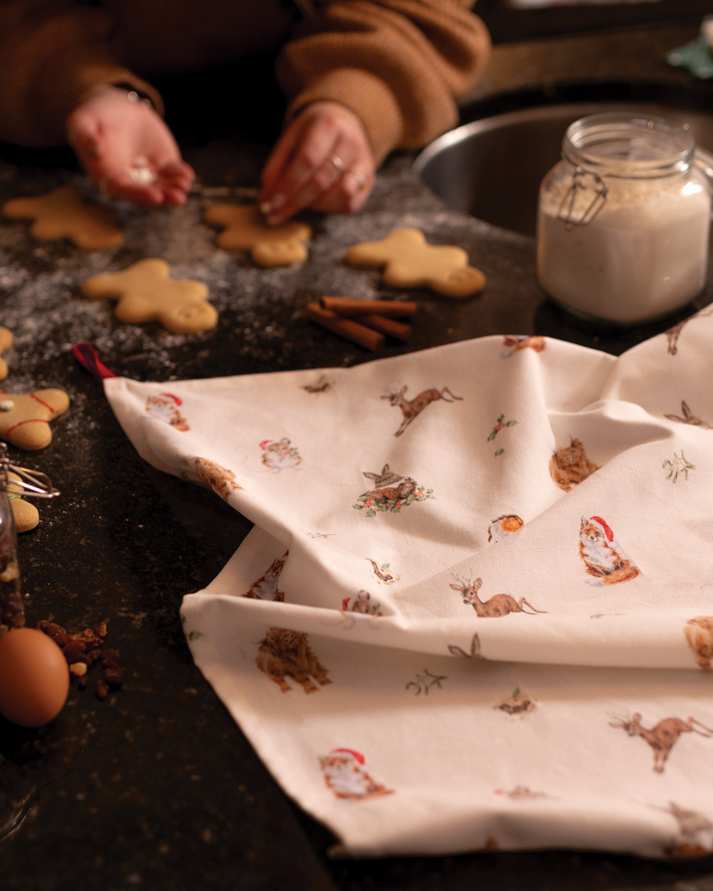 Eine Person verziert Plätzchen auf einer dunklen Küchenarbeitsfläche, daneben liegt das weihnachtliche Woodlanders Geschirrtuch von Wrendale Designs mit Tiermotiven.