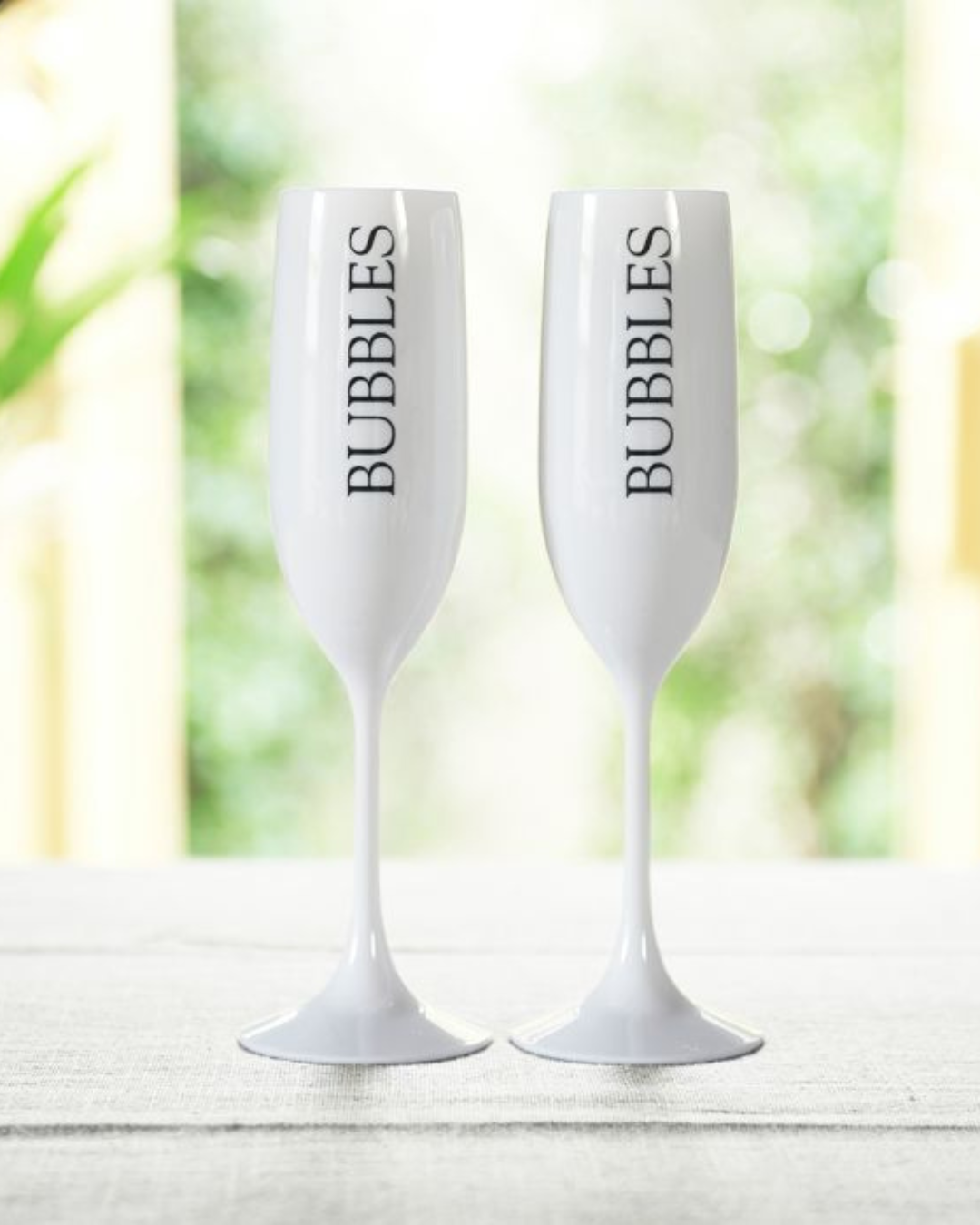 Zwei weiße Bubbles Champagnergläser von Rivièra Maison mit schwarzem Schriftzug ‚BUBBLES‘, auf einem hellen Tisch mit grünem Hintergrund dekoriert.