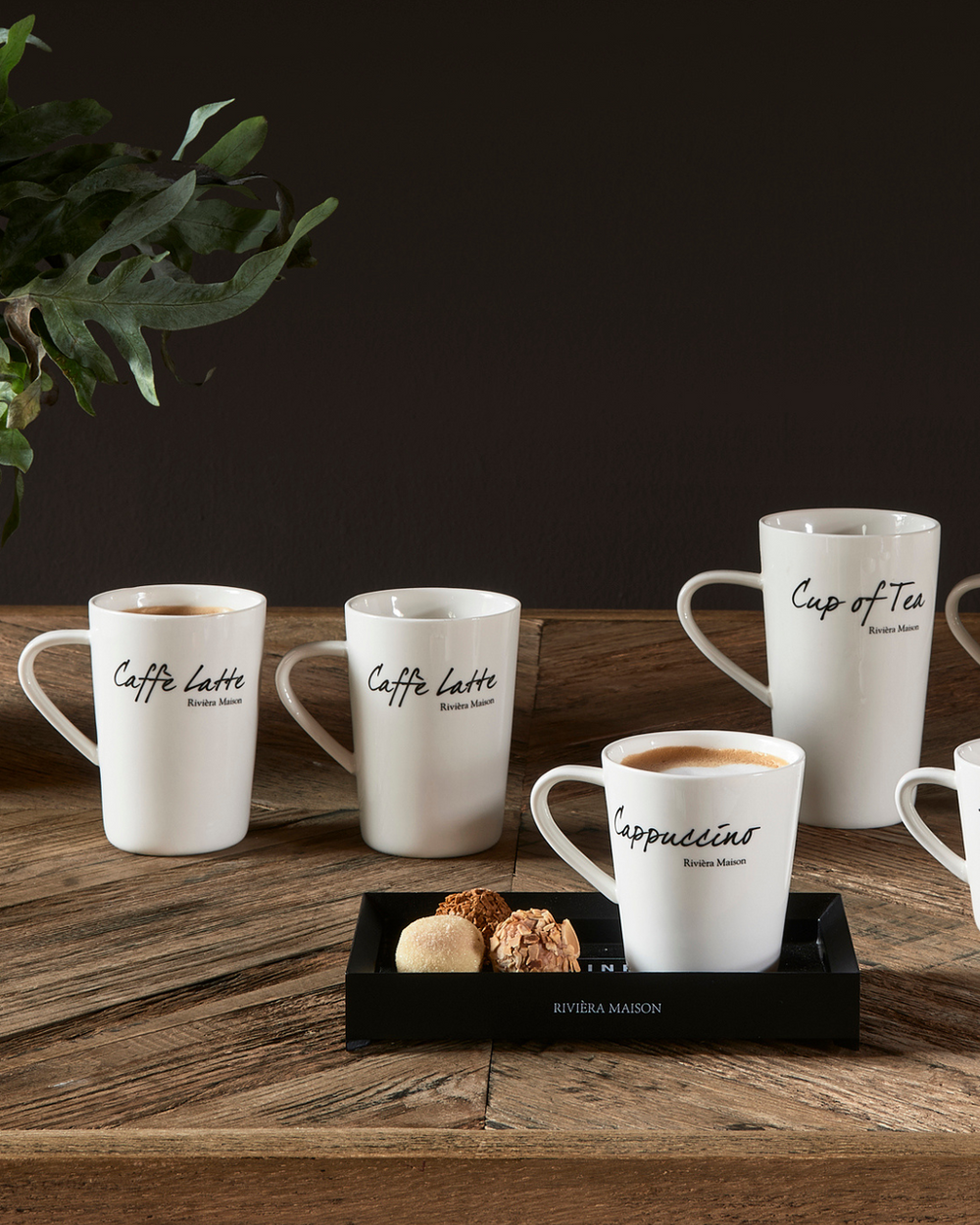 Dekorierte Classic Tassen von Rivièra Maison, mehrere weiße Porzellantassen mit schwarzer Schrift auf einem Holztisch arrangiert, teilweise gefüllt, stimmungsvolle Szene.