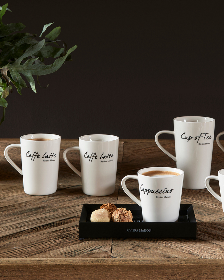 Dekorierte Classic Tassen von Rivièra Maison, mehrere weiße Porzellantassen mit schwarzer Schrift auf einem Holztisch arrangiert, teilweise gefüllt, stimmungsvolle Szene.