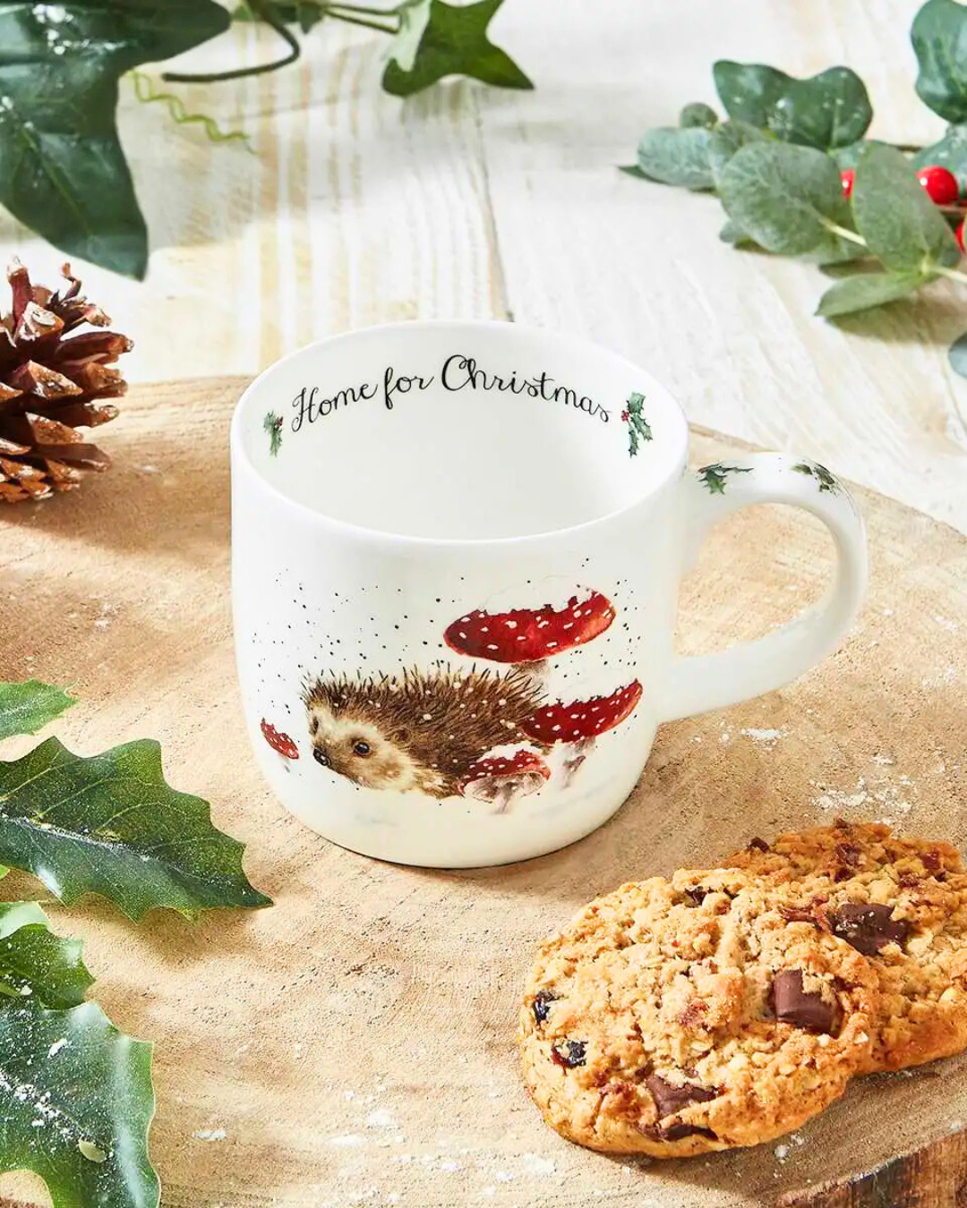 Dekorierte Szene mit der Igel-Tasse „Home for Christmas“ auf einem Holztisch mit Weihnachtsgebäck und Tannenzapfen.