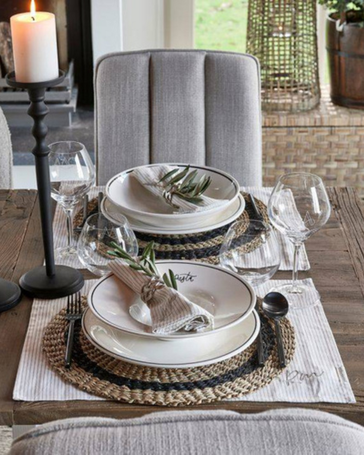 Eleganter Esstisch, gedeckt mit dem 2er Tischset RM Identity von Rivièra Maison. Auf dem gestreiften Platzset liegen weiße Teller mit schwarzen Rändern, Stoffservietten mit Olivenzweigen und Glasgeschirr. Daneben ein schwarzer Kerzenhalter mit brennender Kerze – stilvolle Tischdekoration im modernen Landhausstil.
