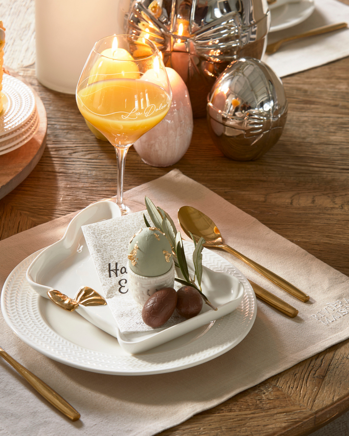 Festlich gedeckter Tisch mit dem klassischen RM Classic Placemat von Rivièra Maison in Beige, kombiniert mit weißen Tellern, goldfarbenem Besteck und stilvoller Osterdeko.