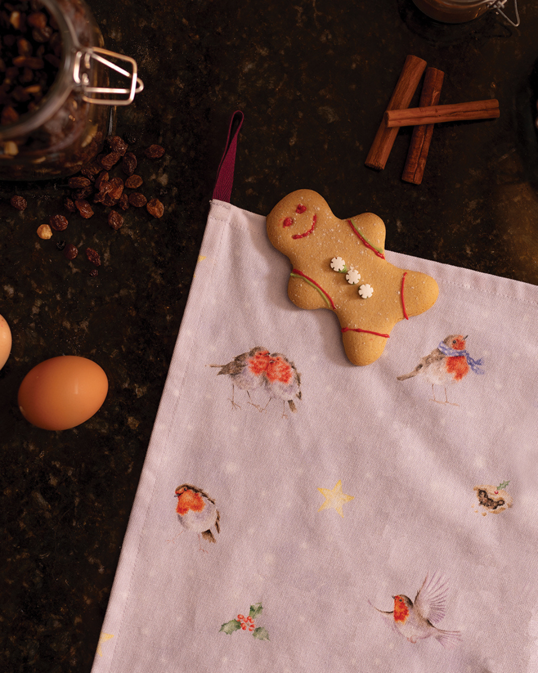 Das Rotkehlchen Geschirrtuch von Wrendale Designs liegt auf einer dunklen Küchenarbeitsfläche. Darauf liegt ein Lebkuchenmann, umgeben von Eiern, Rosinen und Zimtstangen.