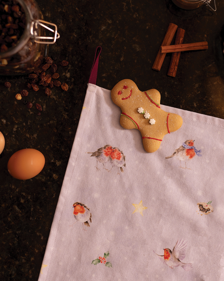 Das Rotkehlchen Geschirrtuch von Wrendale Designs liegt auf einer dunklen Küchenarbeitsfläche. Darauf liegt ein Lebkuchenmann, umgeben von Eiern, Rosinen und Zimtstangen.