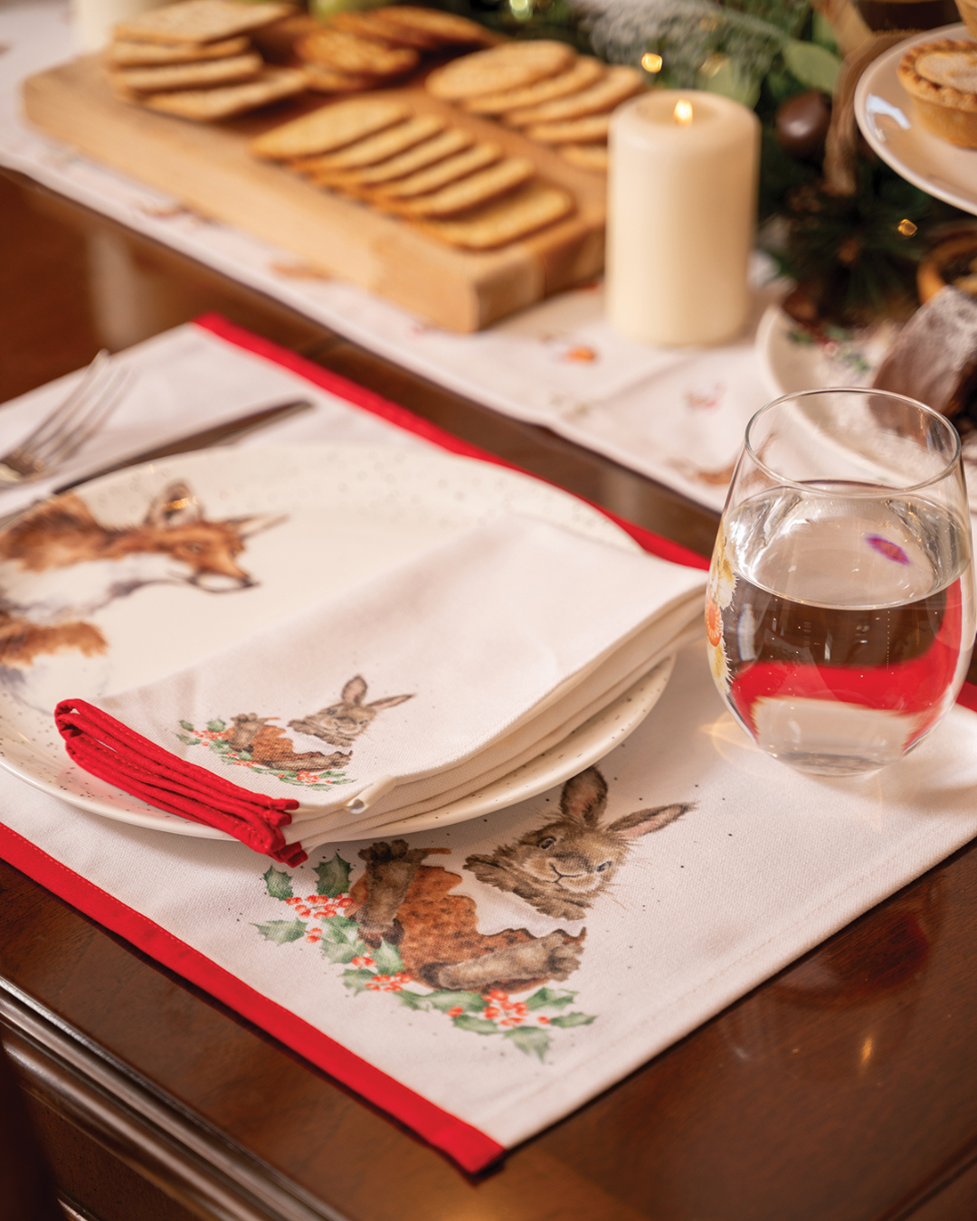 Ein weihnachtlich gedeckter Tisch mit dem Tischset „Winter Woodland“ von Wrendale Designs. Im Vordergrund ist das Hasenmotiv mit rotem Rand zu sehen, dazu Teller, Besteck, ein Glas und eine brennende Kerze für eine gemütliche Winteratmosphäre.