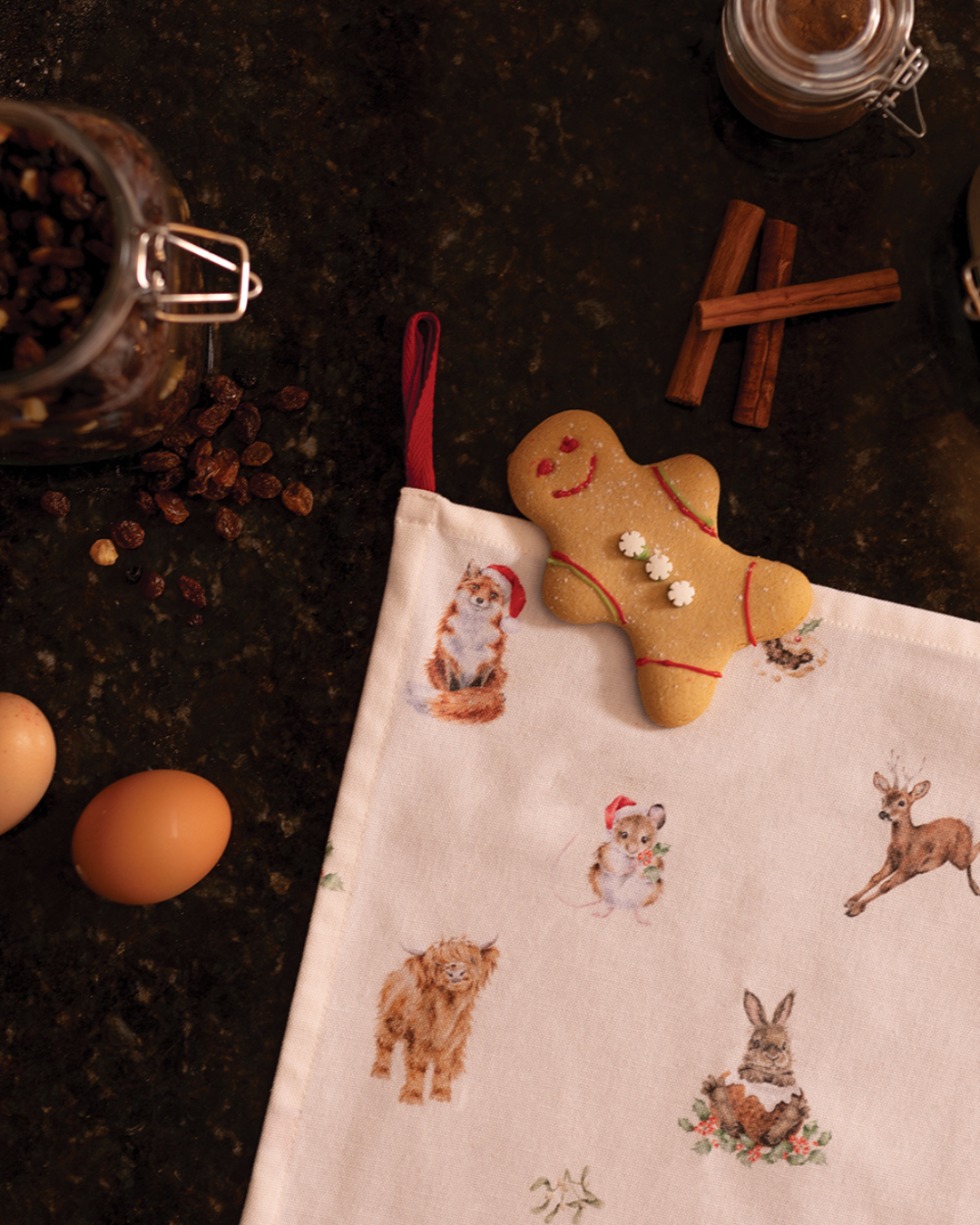 Detailansicht des Woodlanders Geschirrtuchs auf dunklem Untergrund, darauf liegt ein Lebkuchenmann, daneben sind Eier, Rosinen und Zimtstangen zu sehen.