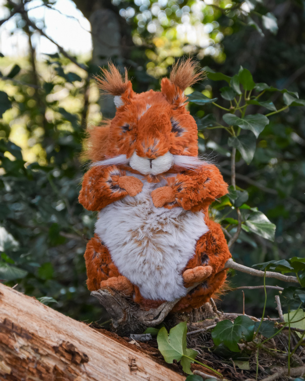 Fern das Eichhörnchen Kuscheltier von Wrendale Designs sitzt auf einem Baumstamm im Wald.