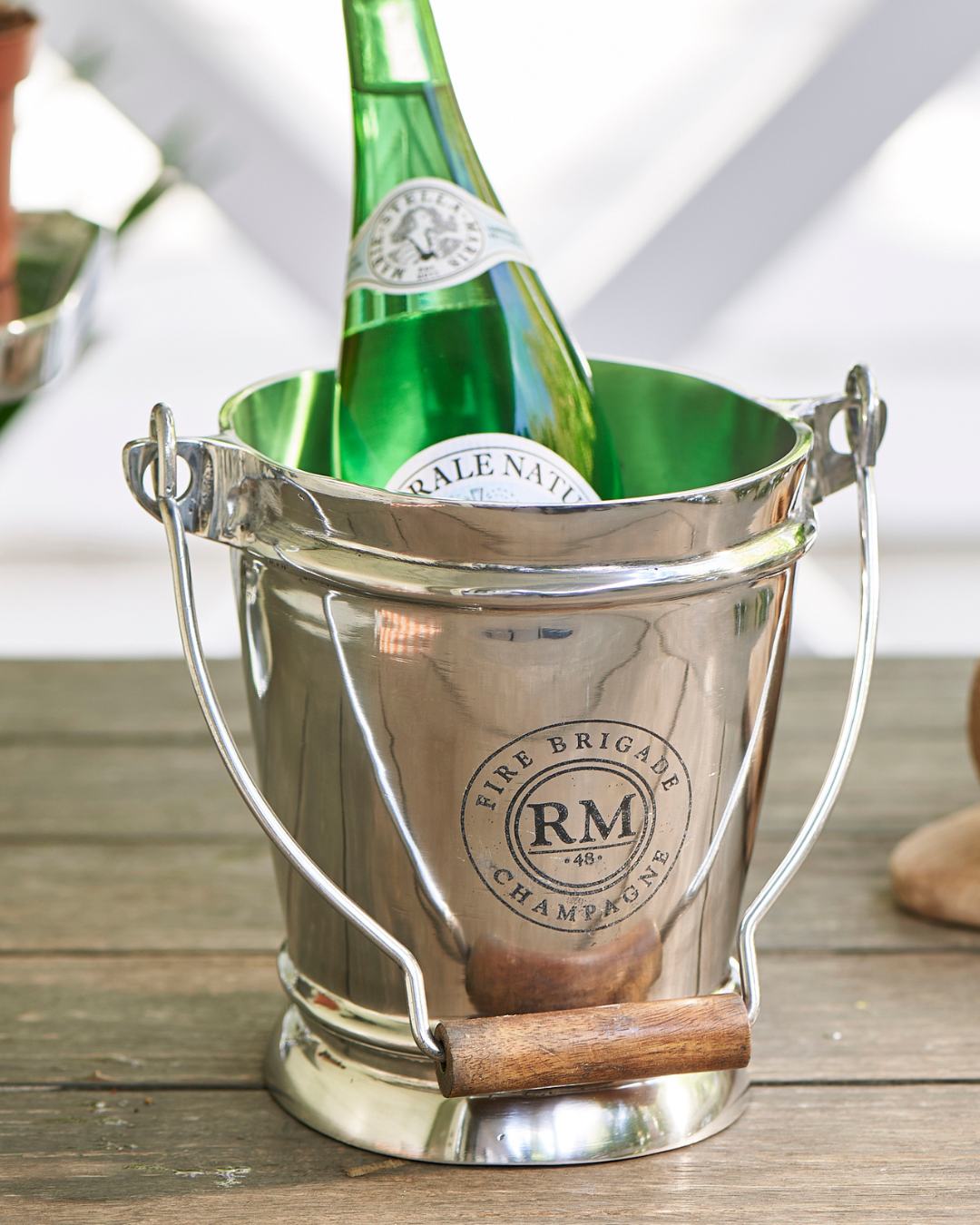 Ein glänzender Fire Brigade Weinkühler von Rivièra Maison mit Holzgriff, gefüllt mit einer grünen Glasflasche, auf einem Holztisch im Freien platziert.