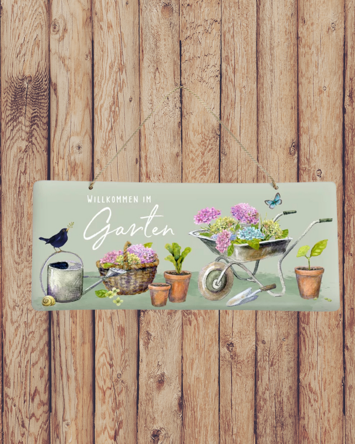 Dekoratives Gartenschild aus Metall mit blumigem Motiv von Daniela Drescher. Hortensien, Schubkarre, Gießkanne und Schmetterling schmücken das liebevolle Design mit Aufschrift „Willkommen im Garten“ an einer Holzwand.