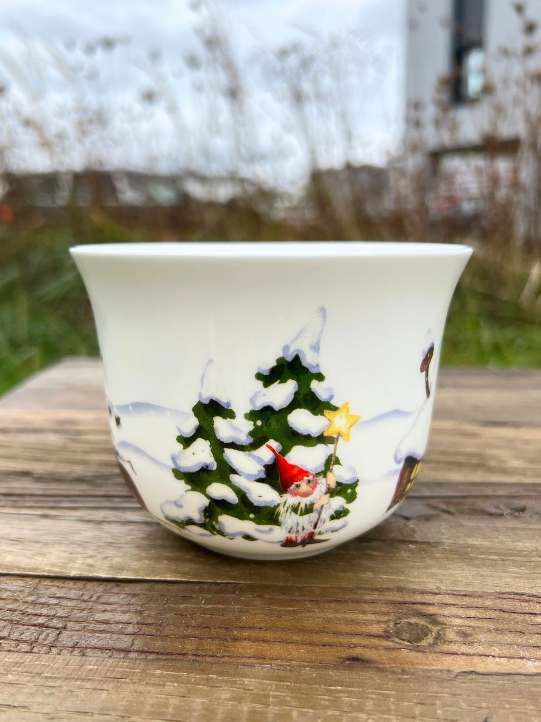 Grätz Verlag Tasse aus Porzellan mit Weihnachtswichtel Motiv auf braunem Tisch in einem Garten.