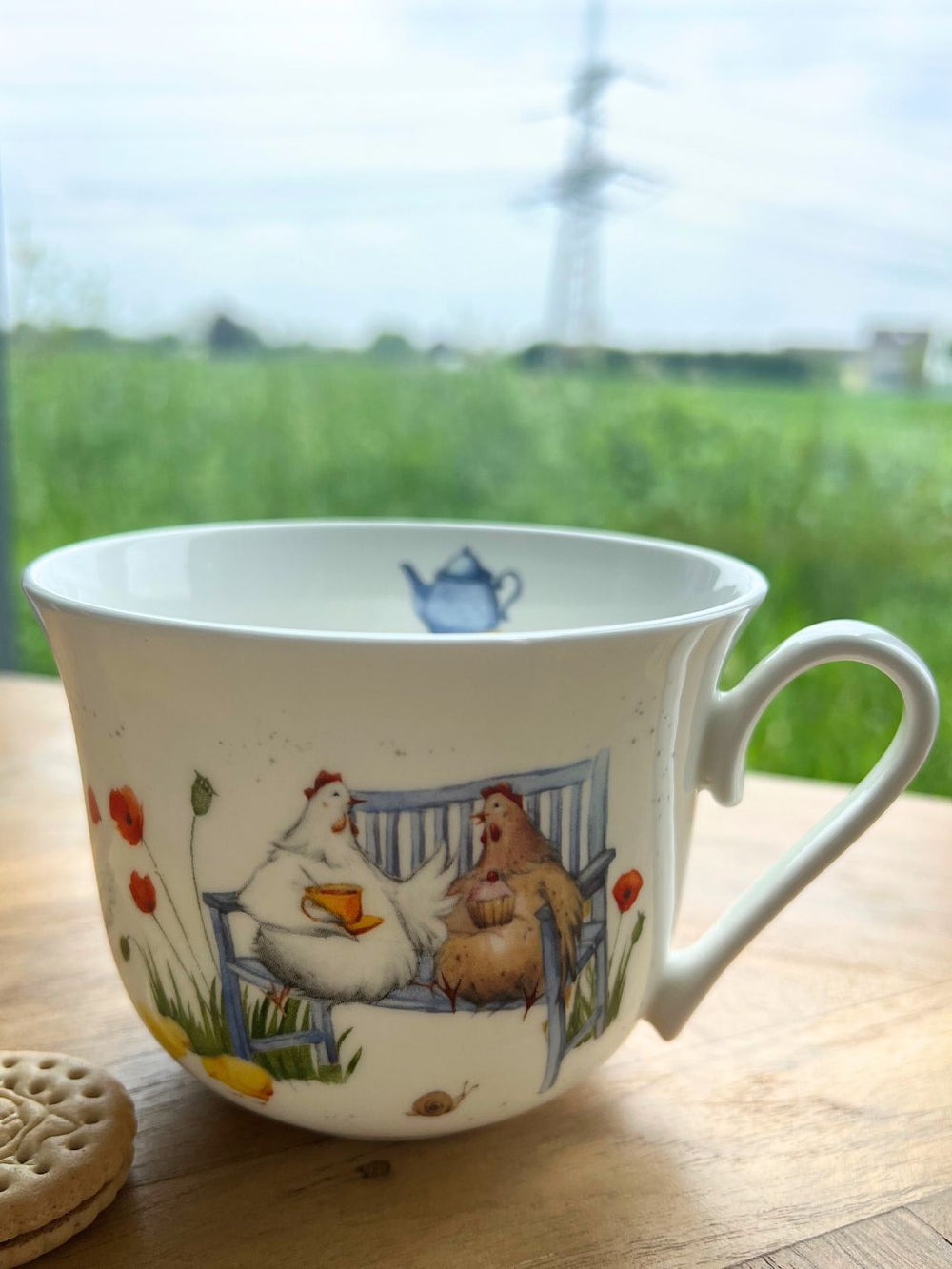 Die Große Hühner Tasse vom Grätz Verlag steht auf einem braunen Holztisch vor grüner Wiese neben einem Keks.
