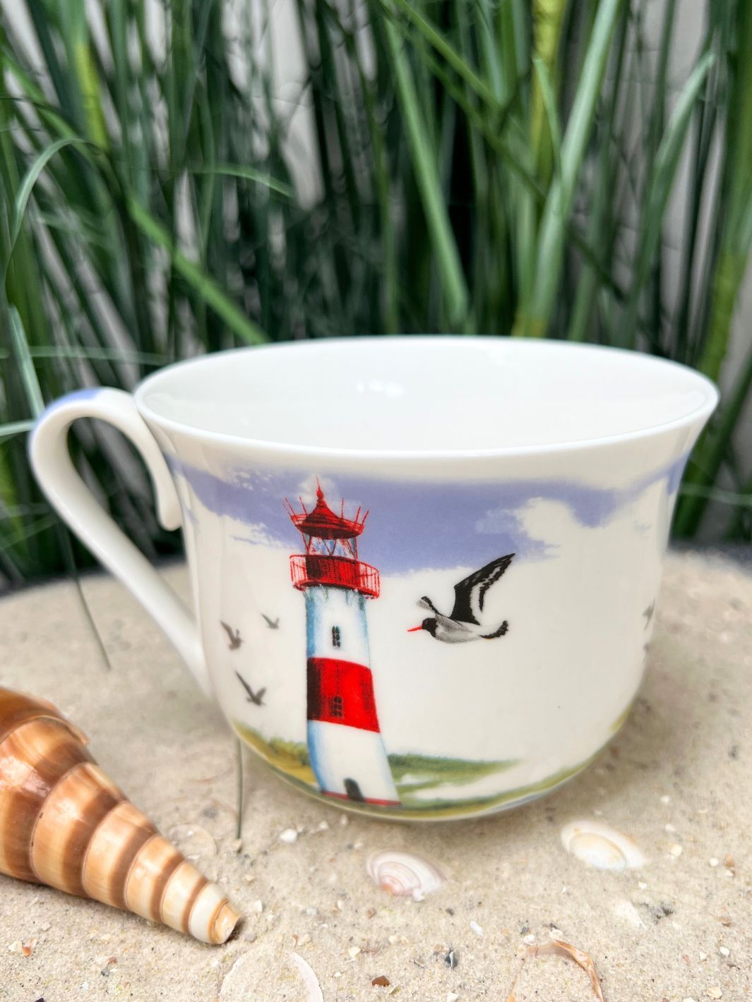Leuchtturm Lieblingstasse vom Grätz Verlag auf einem Tisch mit Sand und Muscheln.