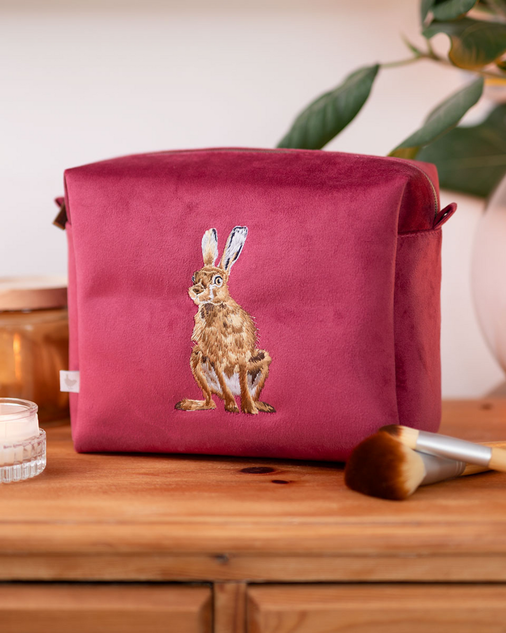 Große weinrote Kosmetiktasche mit gesticktem Hasenmotiv von Wrendale Designs, abgestellt neben Schminke auf einer braunen Holzkommode.