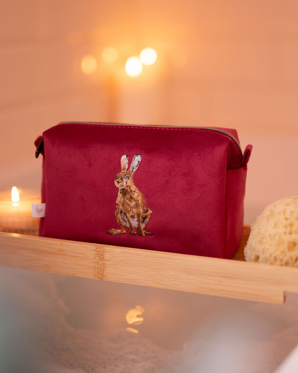 Weinrote Schminktasche mit gesticktem Hasenmotiv von Wrendale Designs auf einem Badewannenbrett, umgeben von Kerzenschein – ideal für Make-up auf Reisen.