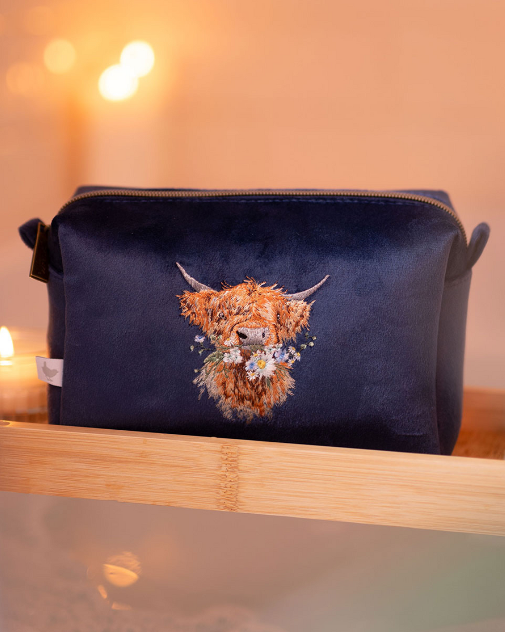 Dunkelblaue Schminktasche von Wrendale Designs mit gestickter Hochlandkuh, platziert auf einem Badewannenbrett mit Kerzenschein im Hintergrund.