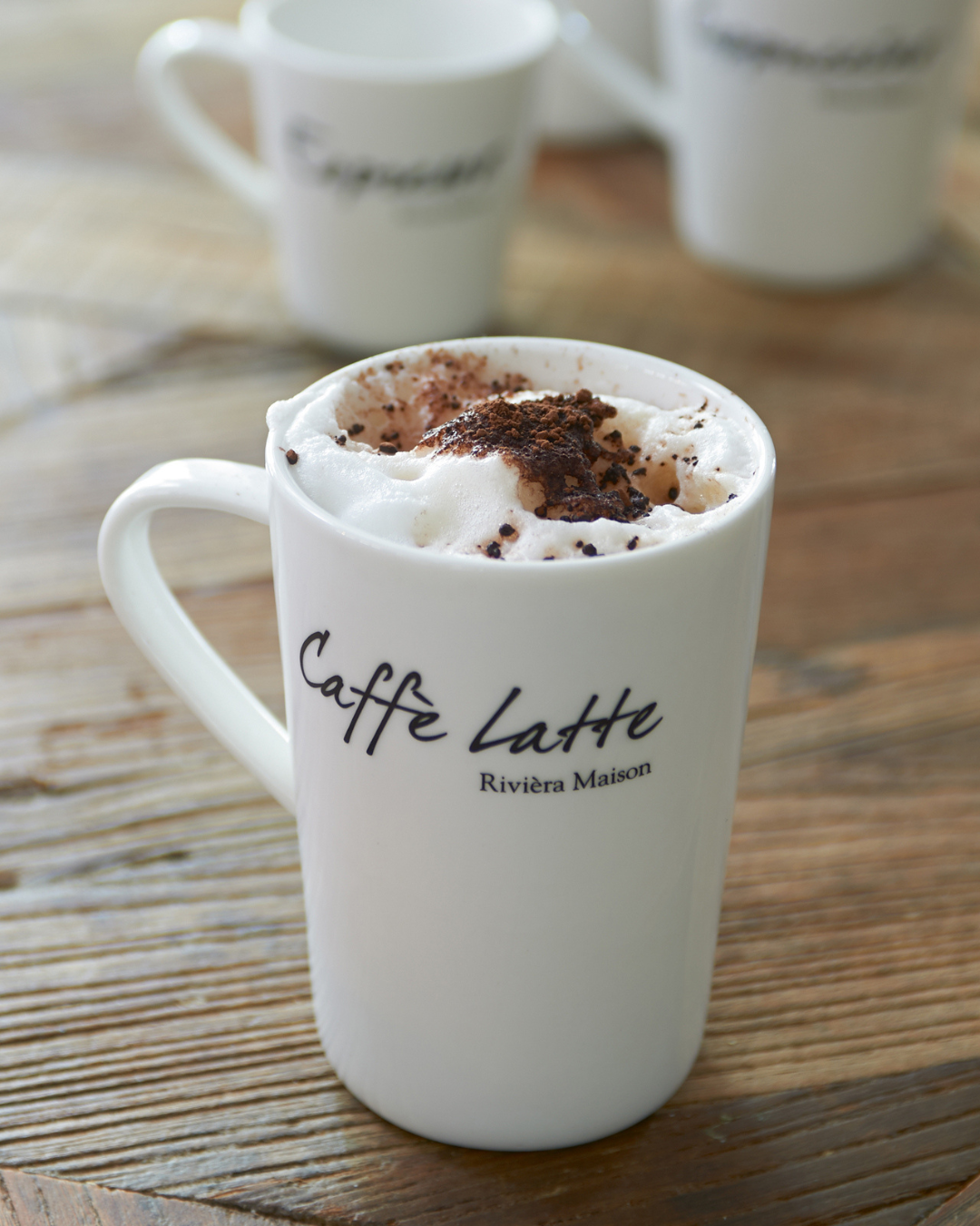 Classic Caffè Latte Tasse von Rivièra Maison, weiße Porzellantasse mit Schriftzug „Caffè Latte“, auf Holztisch stehend und mit Milchschaum sowie Schokoladenstücken dekoriert.