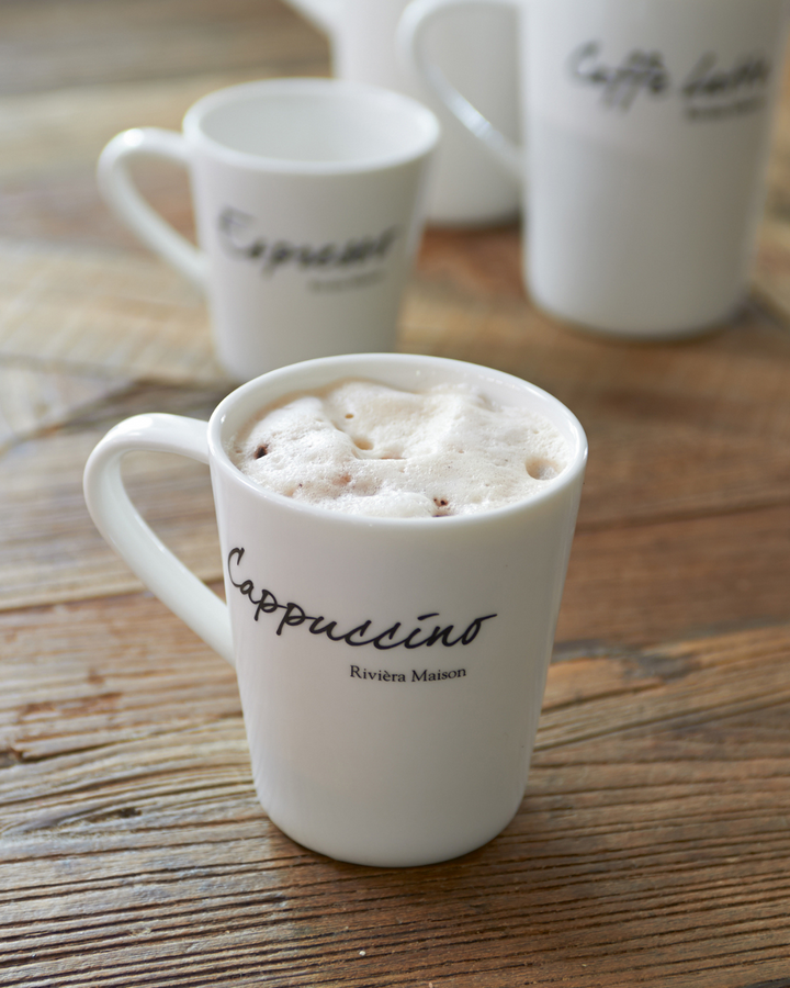 Classic Cappuccino Tasse von Rivièra Maison, weiße Porzellantasse mit Schriftzug „Cappuccino“, auf Holztisch mit cremigem Milchschaum gefüllt.
