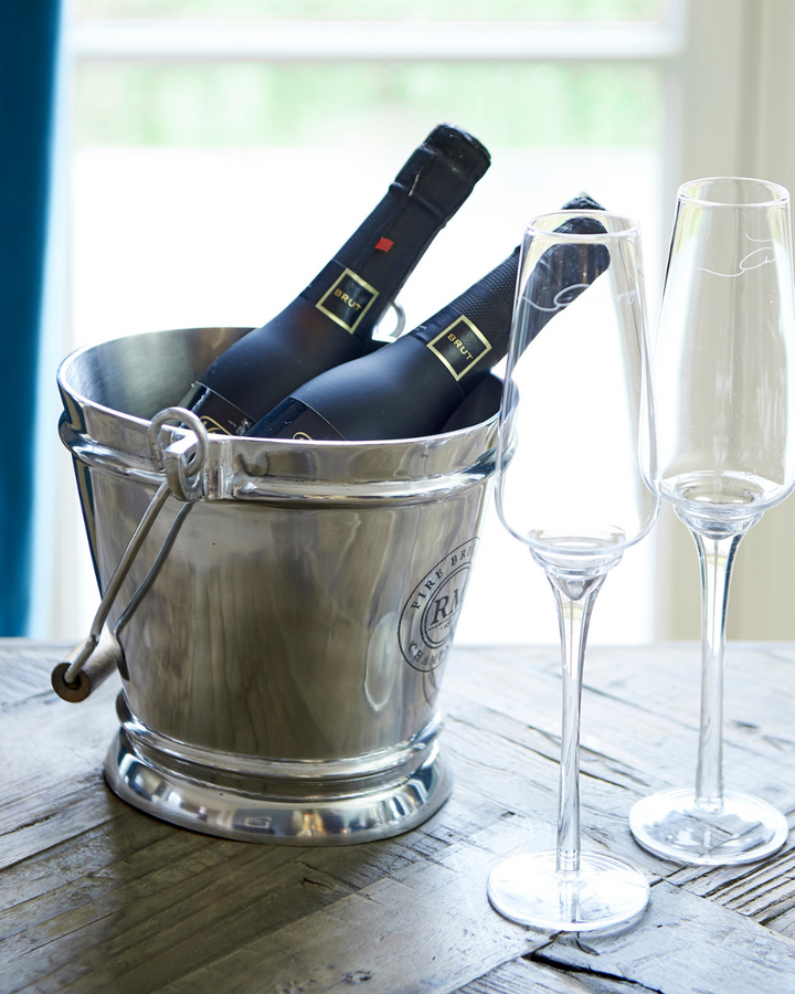 Fire Brigade Weinkühler von Rivièra Maison, gefüllt mit zwei Flaschen Champagner, daneben zwei elegante Sektgläser auf einem Holztisch.