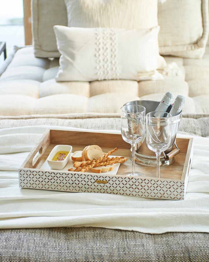 Stilvoll gedecktes Tablett auf einem Bett mit Snacks, Gläsern und dem Fire Brigade Weinkühler von Rivièra Maison mit Sektflaschen.