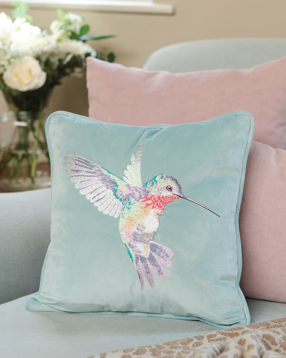 Türkises Kolibri Kissens von Wrendale Designs kombiniert mit rosa Kissen auf einer türkisen Couch.