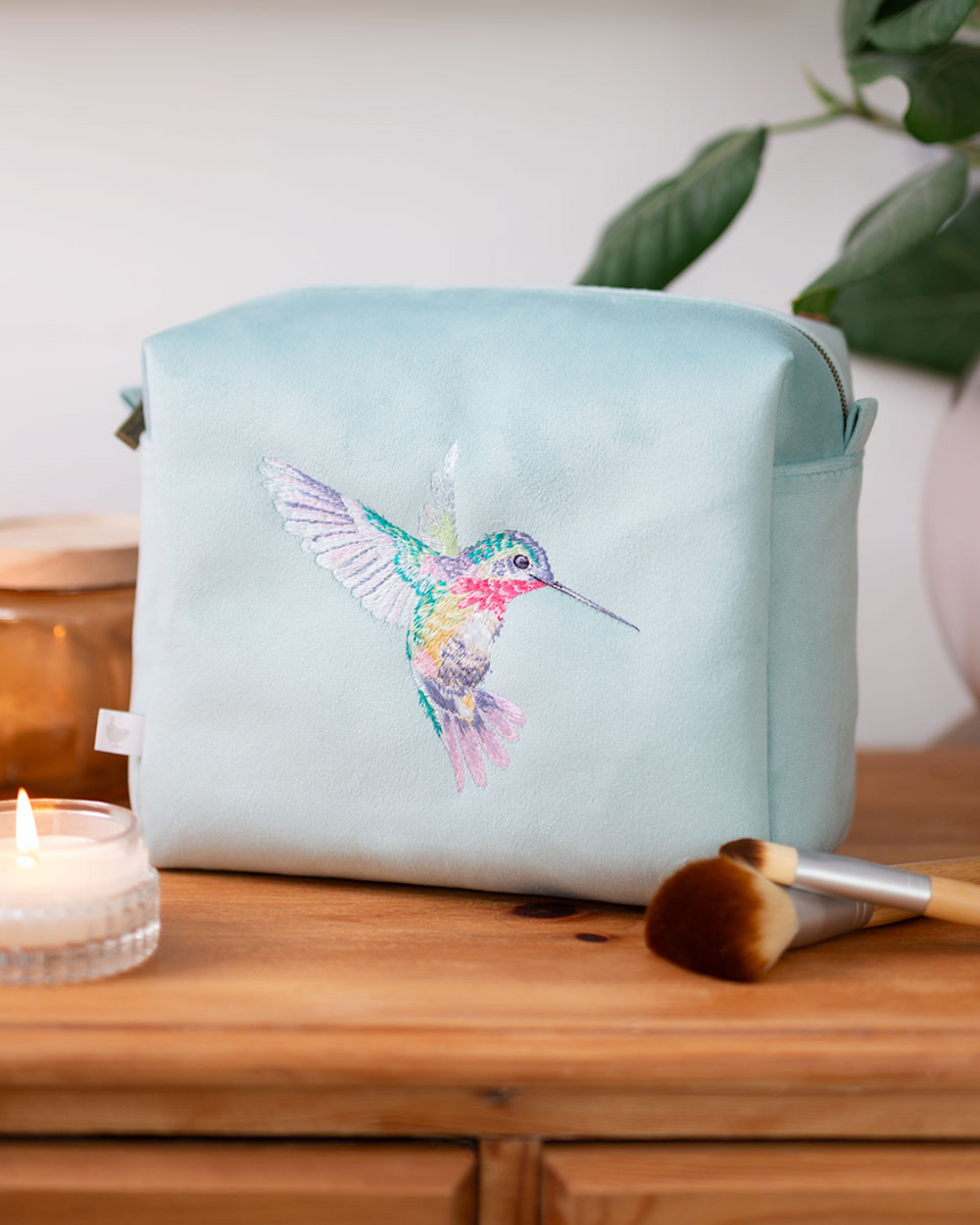 Große türkisfarbene Kosmetiktasche mit gesticktem Kolibri von Wrendale Designs auf einem Holzregal, daneben eine Kerze und Make-up-Pinsel.