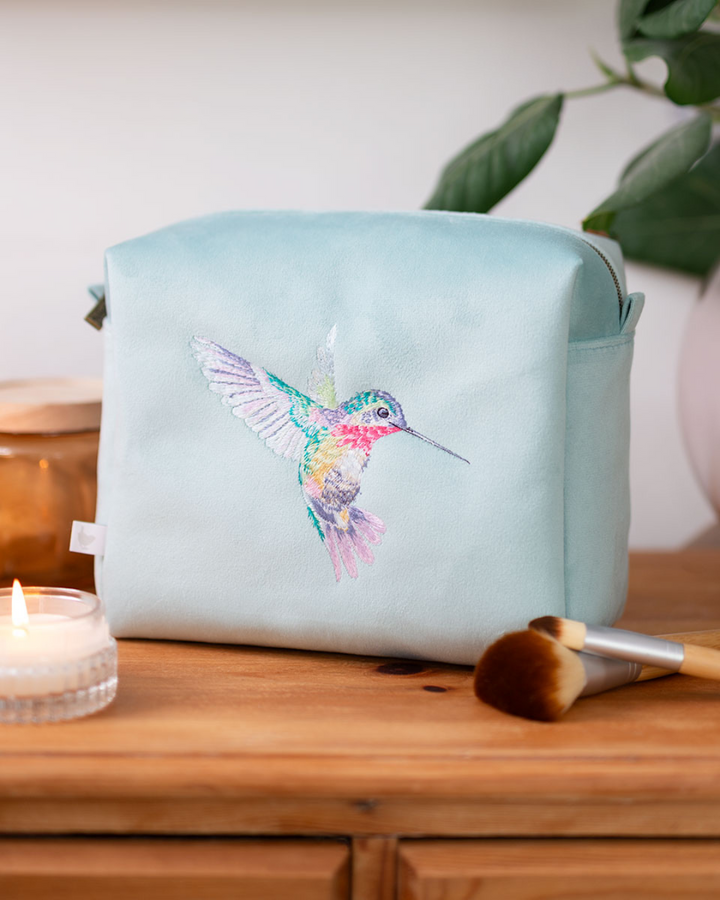 Große türkisfarbene Kosmetiktasche mit gesticktem Kolibri von Wrendale Designs auf einem Holzregal, daneben eine Kerze und Make-up-Pinsel.
