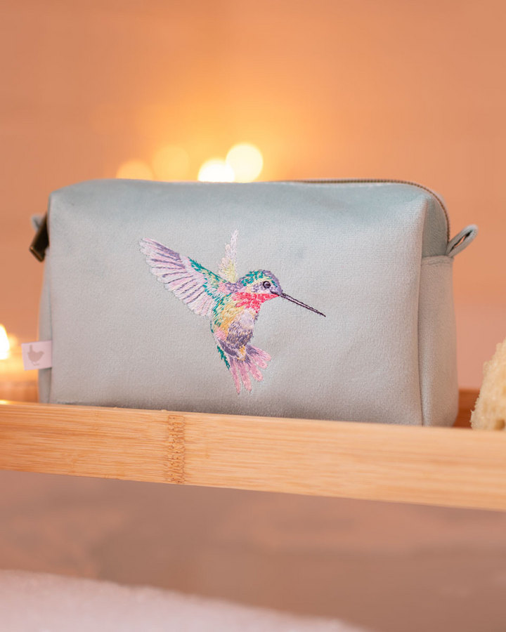Schminktasche mit Kolibri von Wrendale Designs – offen mit Make-up auf einem Wandregal aus Holz arrangiert.