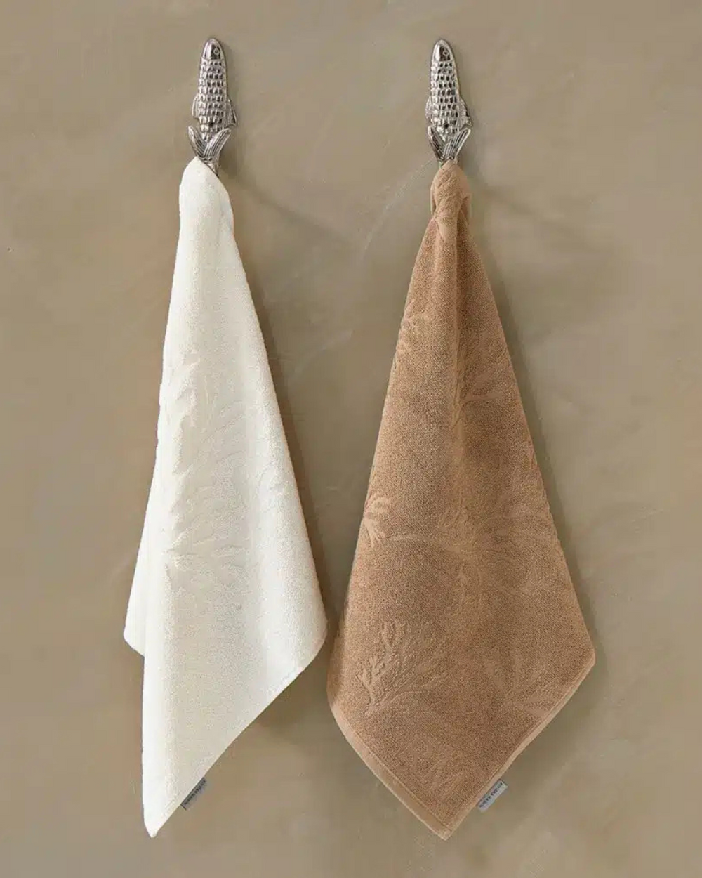 Zwei Küchenhandtücher Coral Reef von Rivièra Maison, eines in Cremeweiß, eines in Sandfarbe, dekorativ nebeneinander an silbernen Wandhaken aufgehängt.