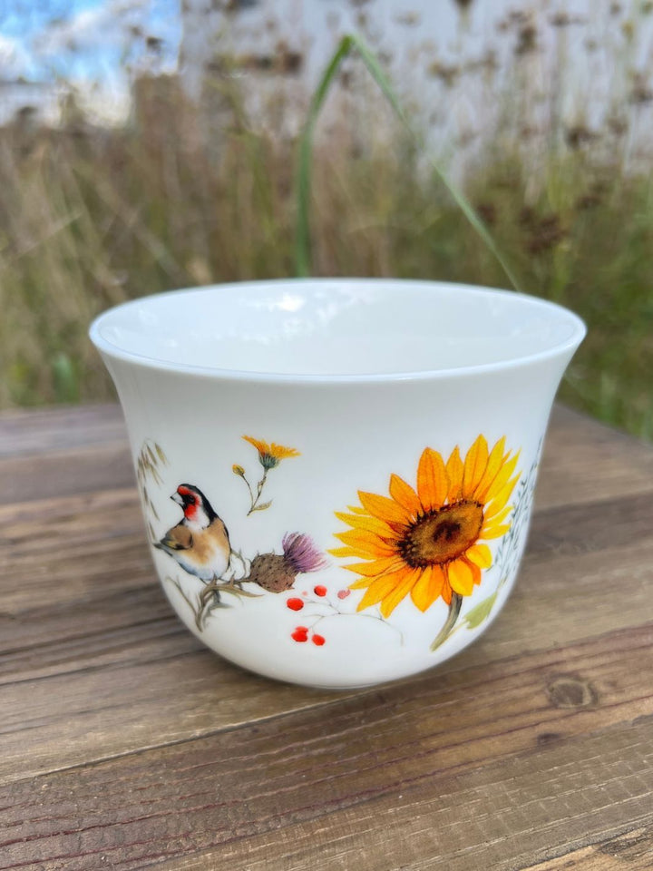 Frontale Ansicht der Lieblingstasse mit Vogel und Sonnenblume drauf vom Grätz Verlag auf einem Holztisch in der grünen Wiese.