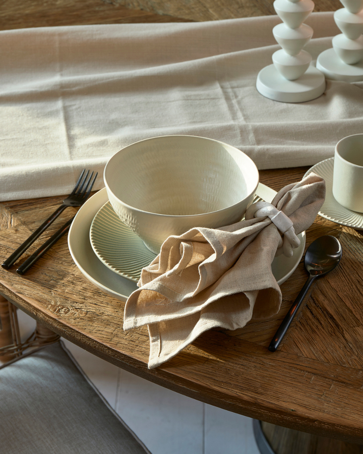 Kombination des Marseille Geschirrs von Rivièra Maison auf einem braunen runden Esstisch mit flaxfarbener Serviette daneben.