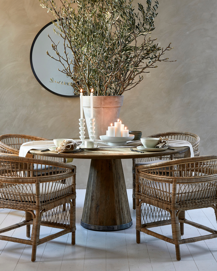 Arrangement des Marseille Geschirrs und weiterer Tischdekoration von Rivièra Maison auf einem runden Holztisch mit vier Stühlen darum, einem großen Olivenbaum im Topf auf dem Tisch und einem Spiegel dahinter an der sandfarbenen Wand.