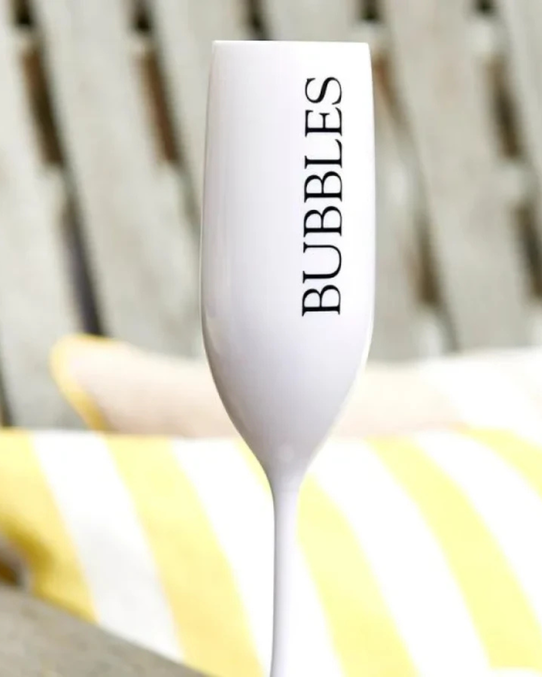 Nahaufnahme des weißen Bubbles Champagnerglases von Rivièra Maison mit schwarzem Schriftzug, im Freien vor Kissen mit gelben und weißen Streifen fotografiert.