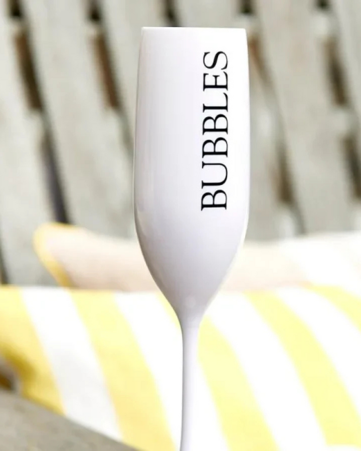 Nahaufnahme des weißen Bubbles Champagnerglases von Rivièra Maison mit schwarzem Schriftzug, im Freien vor Kissen mit gelben und weißen Streifen fotografiert.