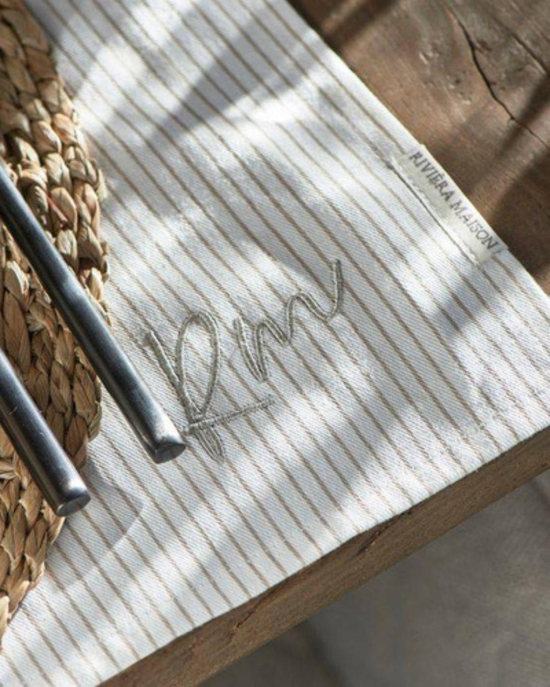 Nahaufnahme des RM Identity Platzsets von Rivièra Maison. Das feine, beige-weiße Streifenmuster mit eingesticktem „RM“-Logo ist gut erkennbar. Daneben liegt Besteck auf einem rustikalen Holztisch – edle Tischwäsche mit dezentem Branding.