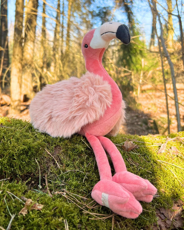 Phoebe der Flamingo als Stofftier von Bukowski sitzt auf einem Baumstamm im Wald.