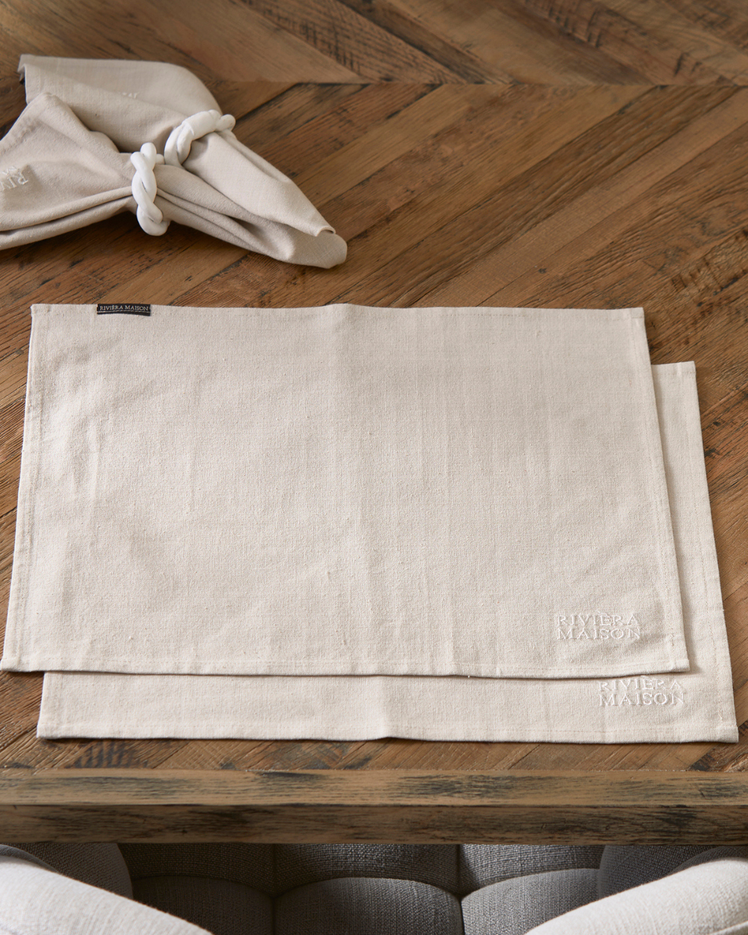Zwei beige RM Classic Placemat Platzsets von Rivièra Maison aus Stoff, mit dezenter Stickerei des Markenlogos in der Ecke.
