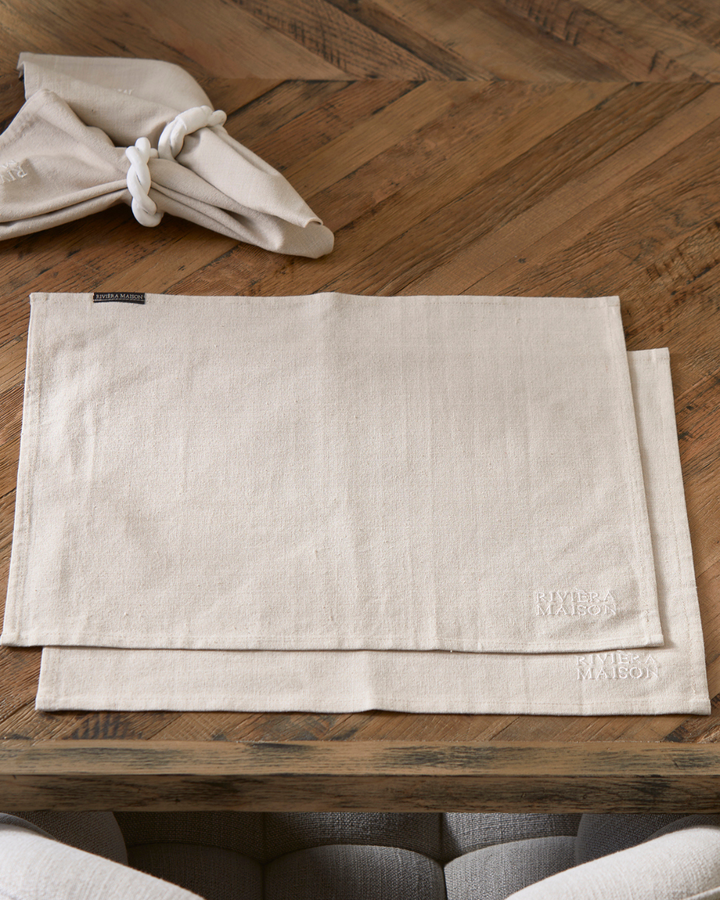 Zwei beige RM Classic Placemat Platzsets von Rivièra Maison aus Stoff, mit dezenter Stickerei des Markenlogos in der Ecke.