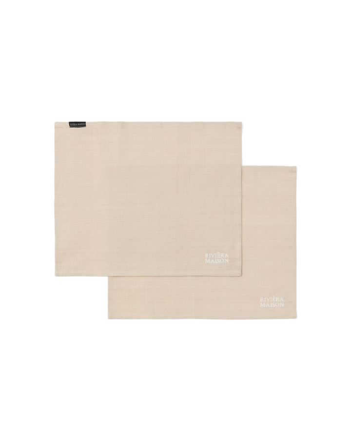 Freigestelltes Bild der RM Classic Placemat Platzsets von Rivièra Maison im 2er-Set, schlicht in Beige mit gesticktem Logo vor weißem Hintergrund.