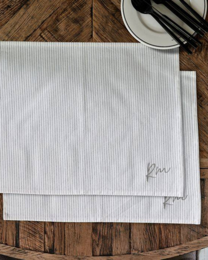 Nahaufnahme des RM Identity Platzsets von Rivièra Maison. Das feine, beige-weiße Streifenmuster mit eingesticktem „RM“-Logo ist gut erkennbar. Daneben liegt Besteck auf einem rustikalen Holztisch – edle Tischwäsche mit dezentem Branding.