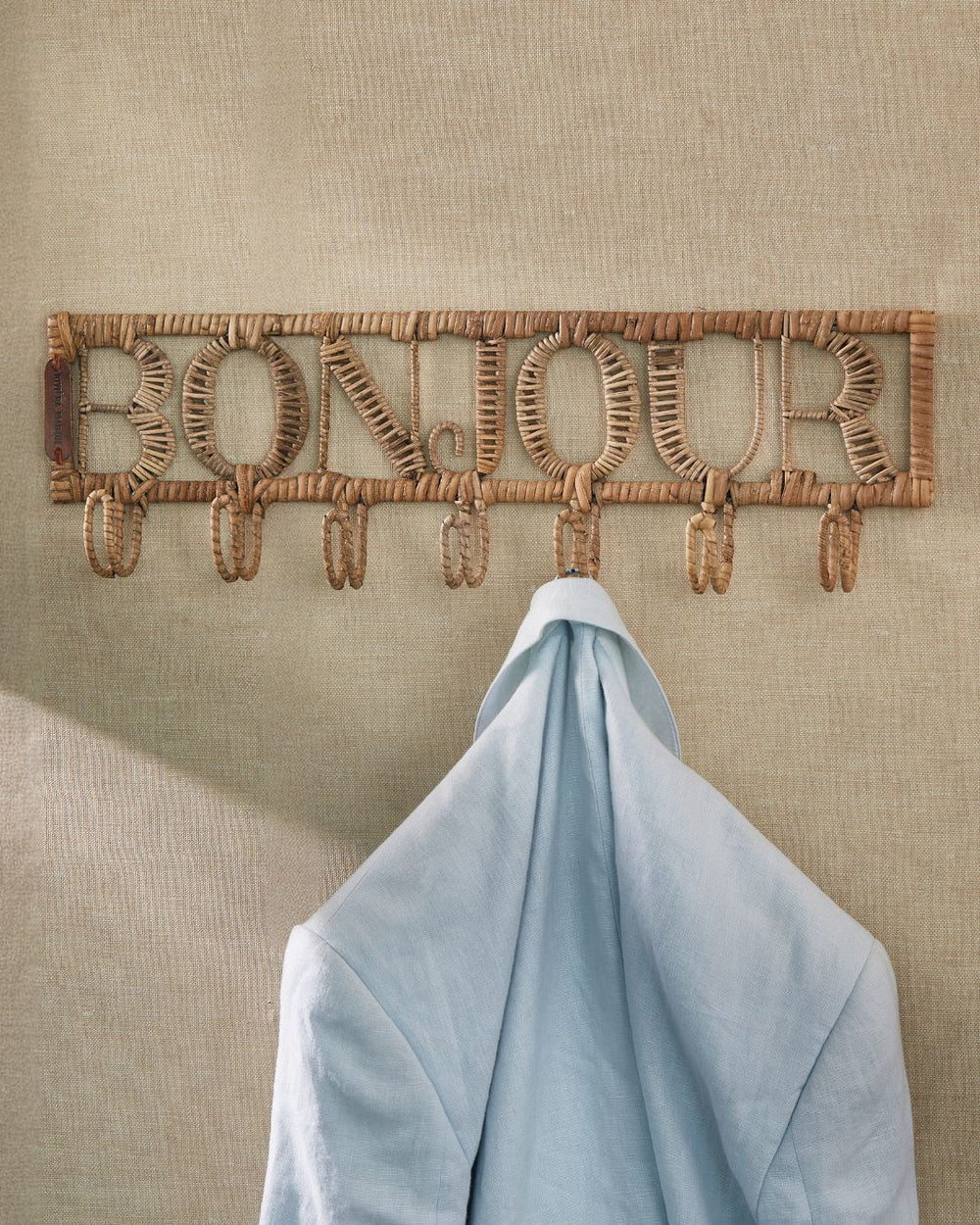Die Bonjour Garderobe von Rivièra Maison ist an einer dunkelgelben Wand angebracht und eine hellblaue Jacke hängt daran.