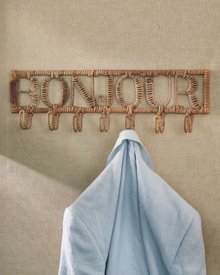 Die Bonjour Garderobe von Rivièra Maison ist an einer dunkelgelben Wand angebracht und eine hellblaue Jacke hängt daran.
