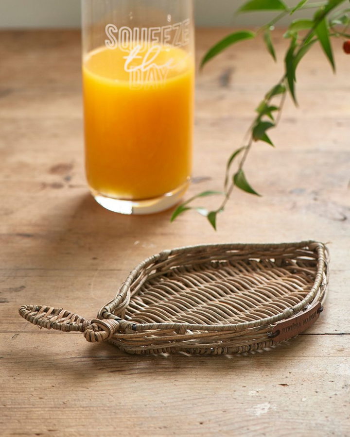 Das Zitronen Tablett aus Rattan von Rivièra Maison liegt auf einem braunen Holztisch neben einer Flasche Orangensaft.