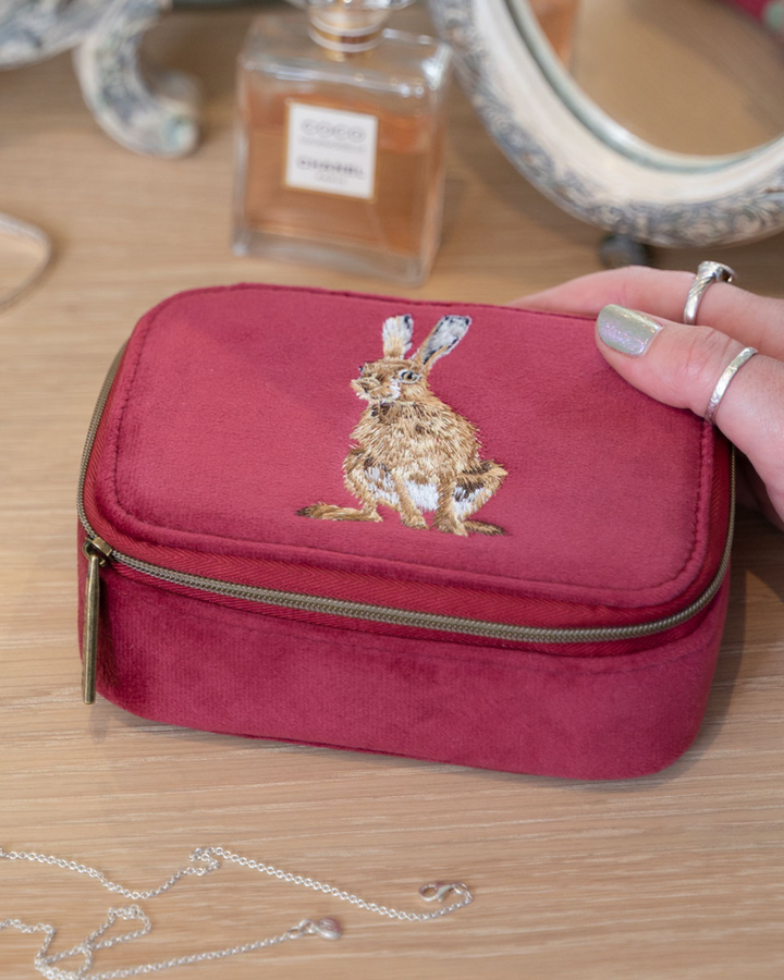 Weinrote Schmucktasche mit Hasenmotiv von Wrendale Designs auf einem Schminktisch neben einem Parfumflakon.