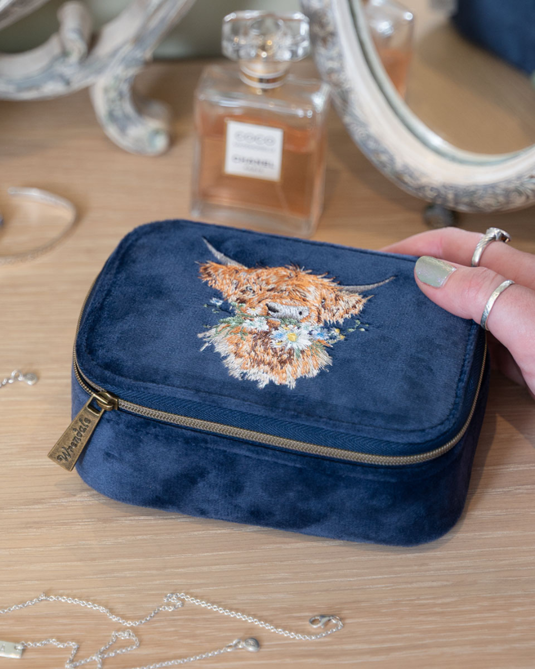 Dunkelblaue Schmucktasche mit Hochlandkuhmotiv von Wrendale Designs auf einem Schminktisch neben einem Parfumflakon.