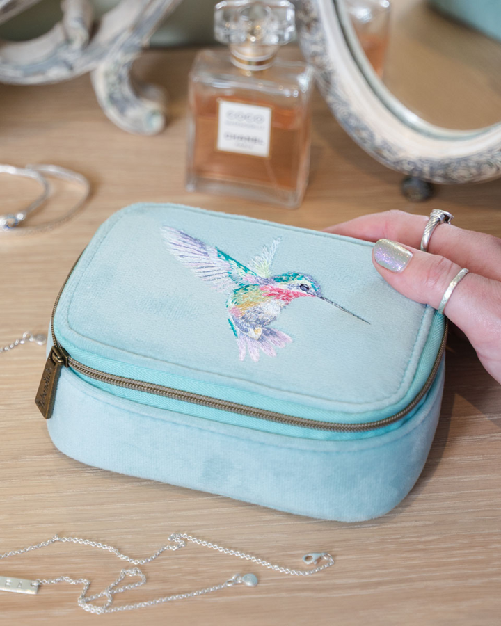 Türkise Schmucktasche mit Kolibrimotiv von Wrendale Designs auf einem Schminktisch neben einem Parfumflakon.
