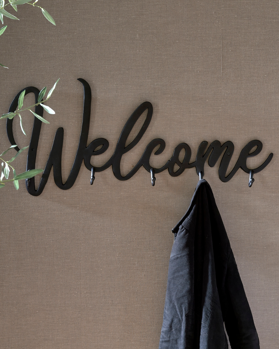 Schwarze „Welcome“-Garderobe mit fünf Haken von Rivièra Maison vor brauner Wand, eine schwarze Jacke hängt daran, Hintergrund neutral.
